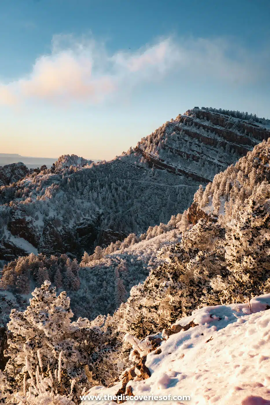 Snow-covered forest and the layered rock ridge of the Sandia Mountains crest under a blue sky with clouds