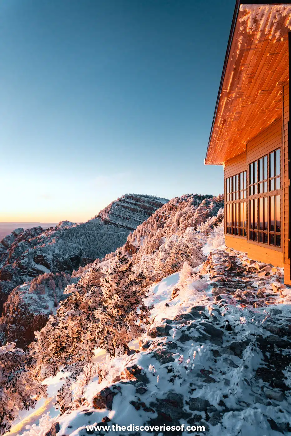 Red-roofed top station of the Sandia Peak Tramway beside snowy rocks and trees in golden evening light