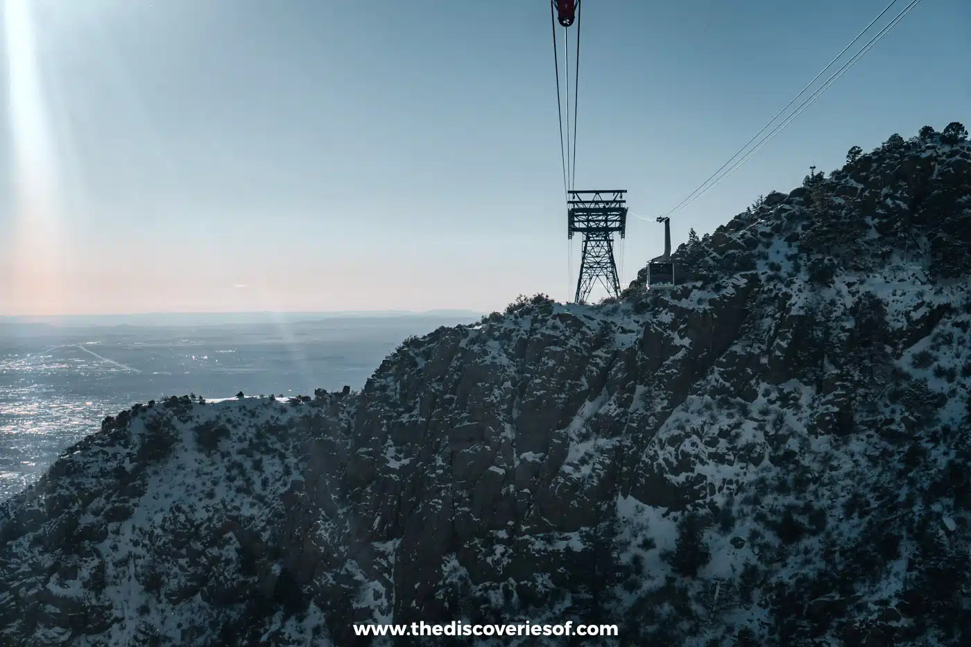 Sandia Peak Tramway car and support tower above snow-dusted ridges, with Albuquerque spread out below in winter sun