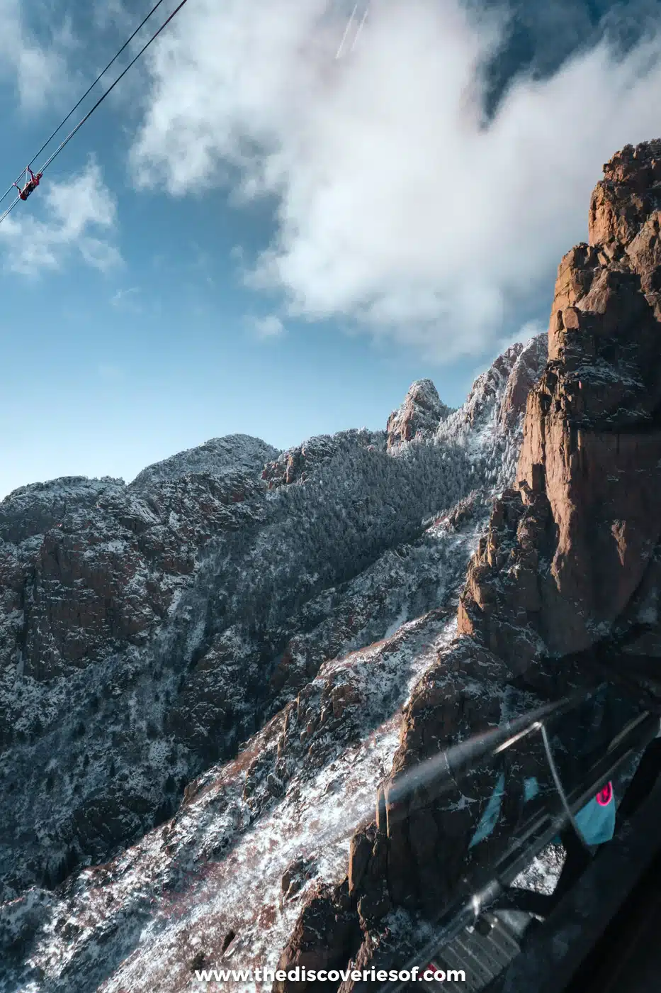 Snow-dusted granite cliffs and a tram cable seen from the cabin of the Sandia Peak Tramway on the way up