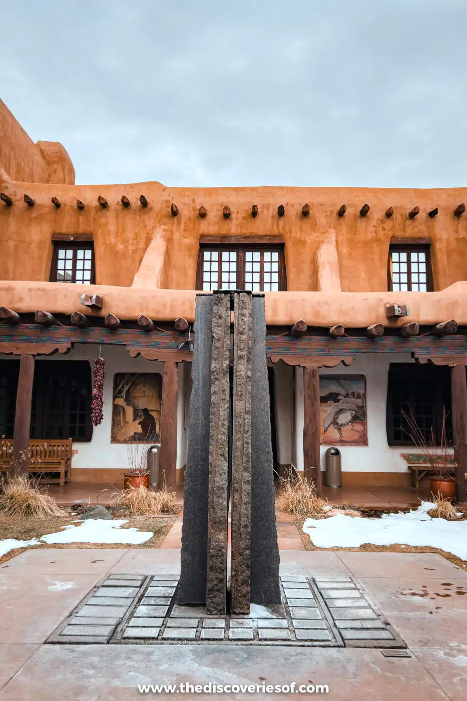 Tall dark sculpture in the snowy adobe courtyard of the New Mexico Museum of Art in Santa Fe