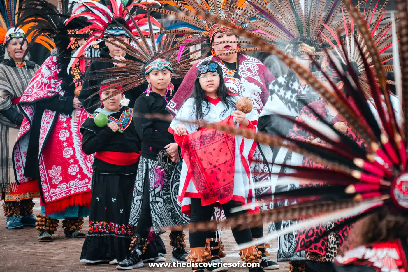 Dancers in red and black regalia with tall feathered headdresses at a gathering on Taos Plaza