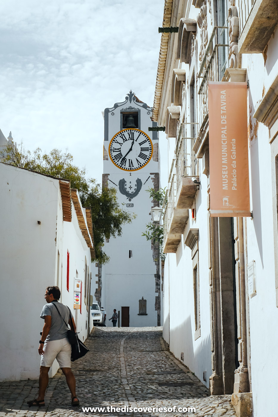 A cobbled lane in Tavira past the Museu Municipal banner on the Palácio da Galeria, leading up to the clock tower of Santa Maria do Castelo