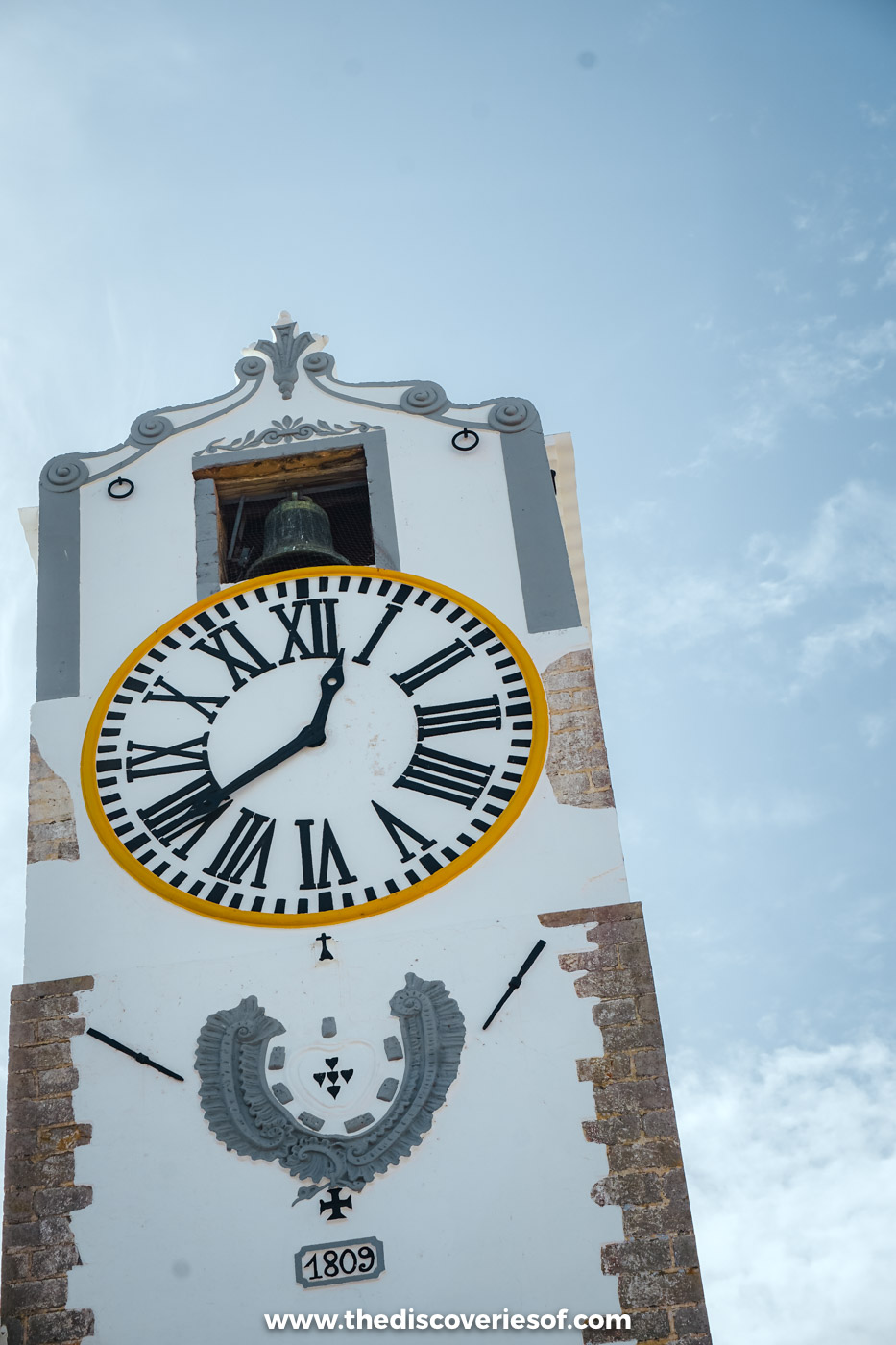 The white clock tower of Santa Maria do Castelo in Tavira, with its bell, Roman numerals and carved coat of arms