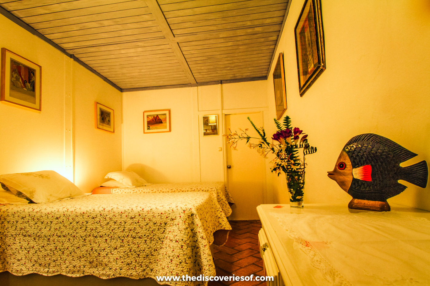Twin beds with floral covers, fresh flowers and a carved wooden fish in a warmly lit yellow room at Quinta do Barranco da Estrada