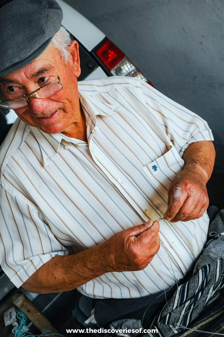 An older fisherman in a flat cap and glasses mending a fishing line by hand in Azenha do Mar