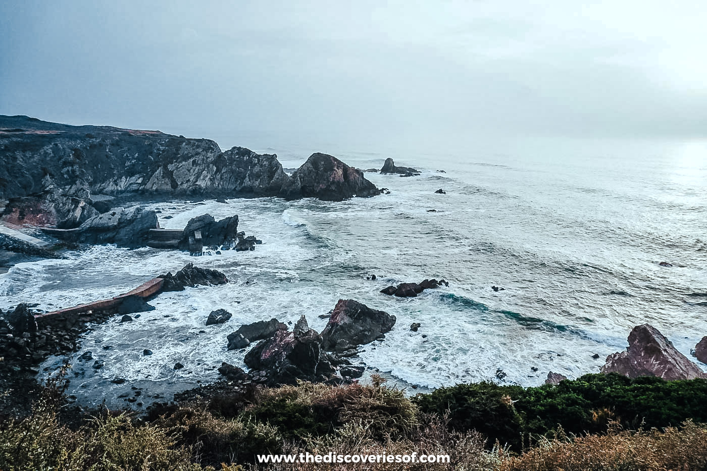 White surf breaking over dark and rust-coloured rocks and old concrete breakwaters in a rocky cove below the clifftop path
