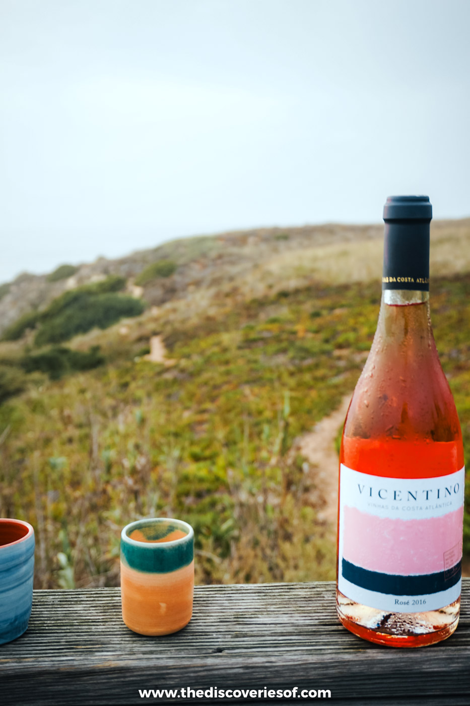 A bottle of Vicentino rosé and two ceramic cups on a wooden boardwalk rail above the Fishermen's Trail near Zambujeira do Mar