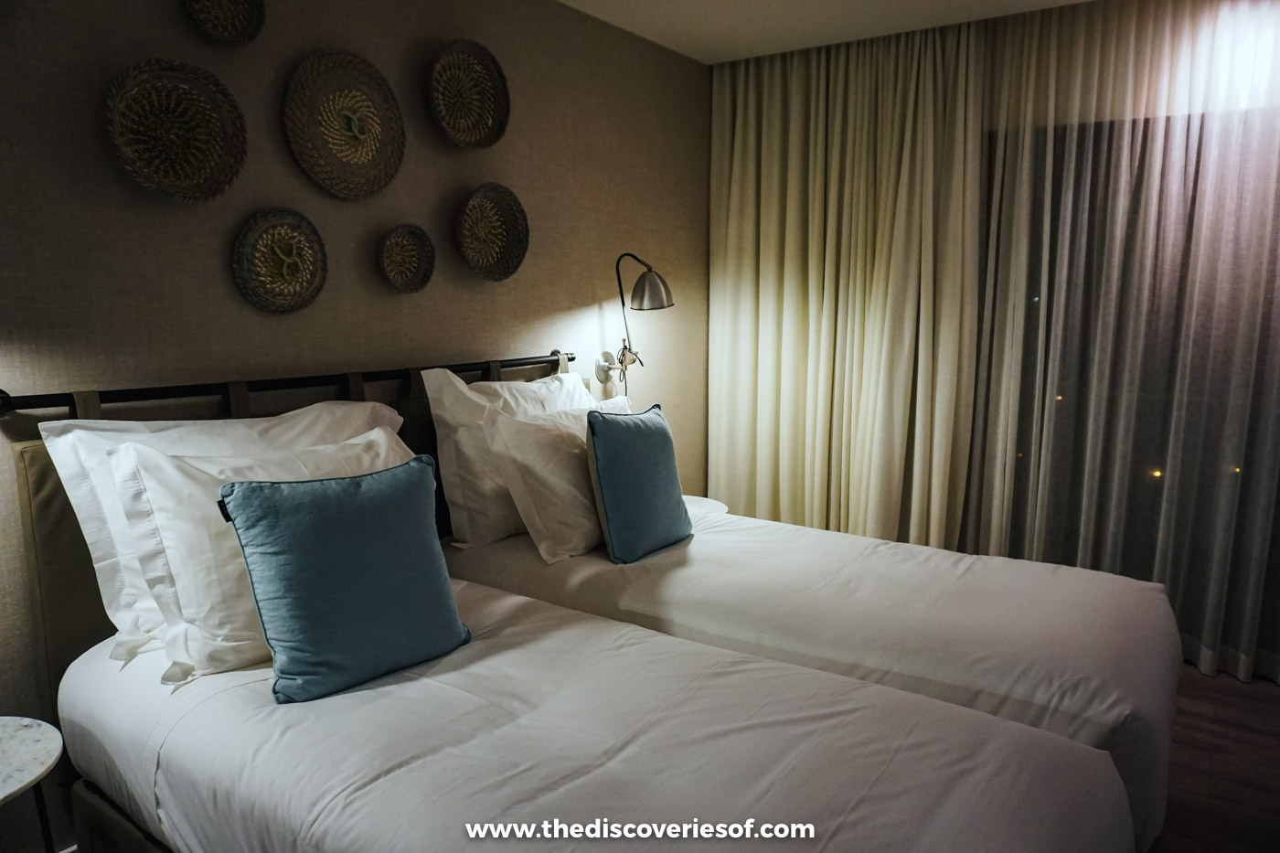 Twin beds with white linen and blue cushions below woven baskets hung on the wall of a bedroom at Octant Santiago