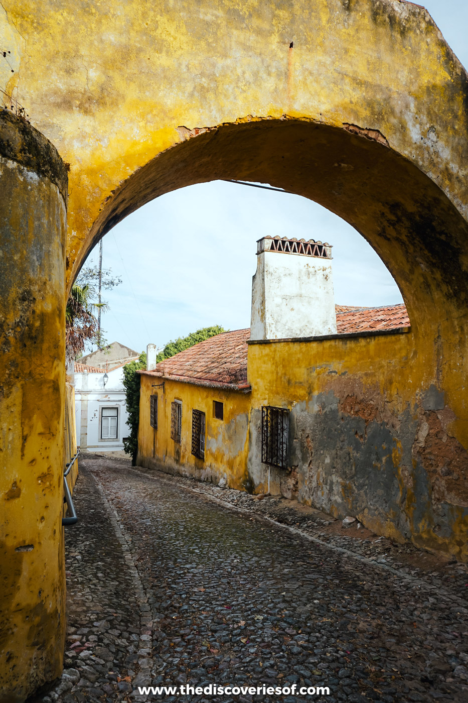 A cobbled lane running under a weathered yellow archway past old yellow houses in the old town of Santiago do Cacém
