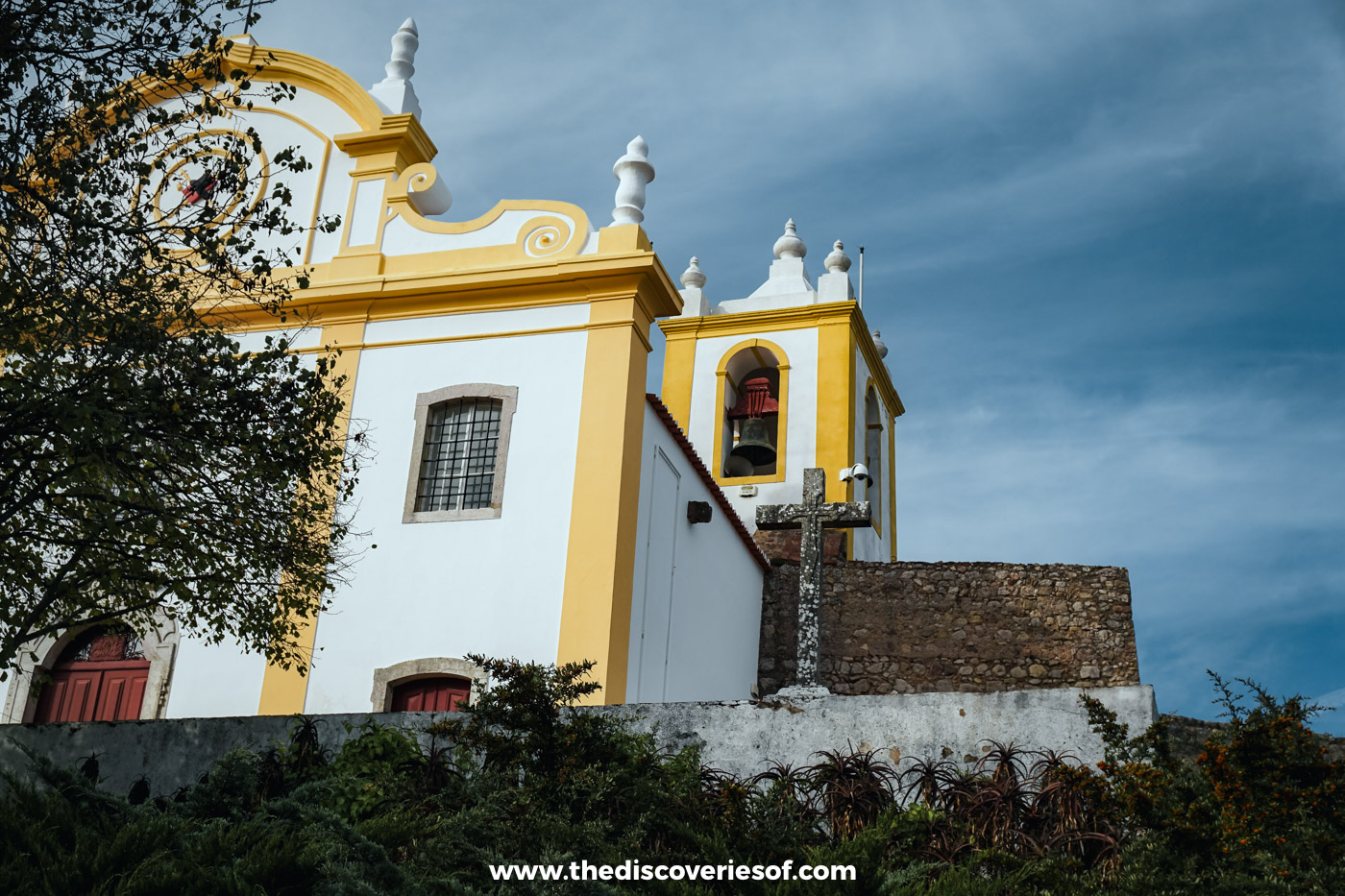 The white and yellow baroque facade and bell tower of a church below a stone wall under a stormy sky in Santiago do Cacém