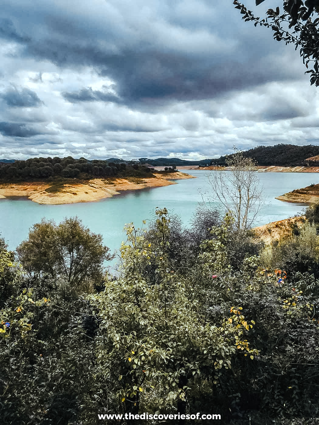 The turquoise water of the Santa Clara reservoir between wooded hills under a cloudy sky in the Alentejo