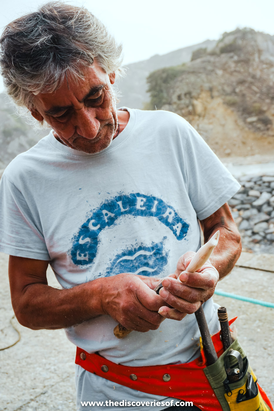 A grey-haired fisherman in a white T-shirt and red waistband working with a wooden tool by the harbour at Azenha do Mar