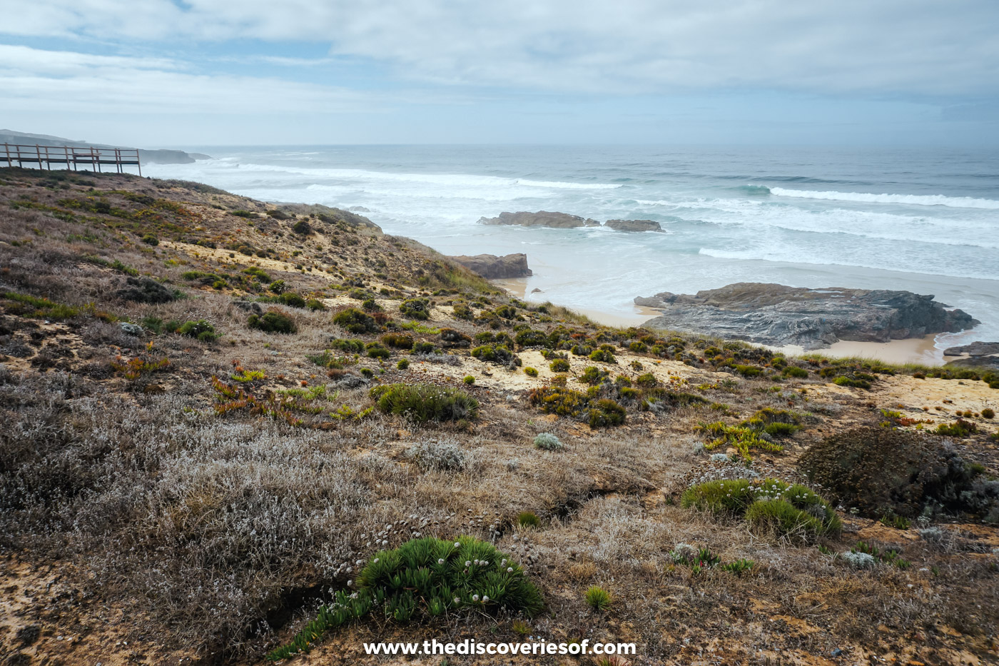 Grey surf breaking on rocks below scrub-covered cliffs and a boardwalk on the Alentejo coast