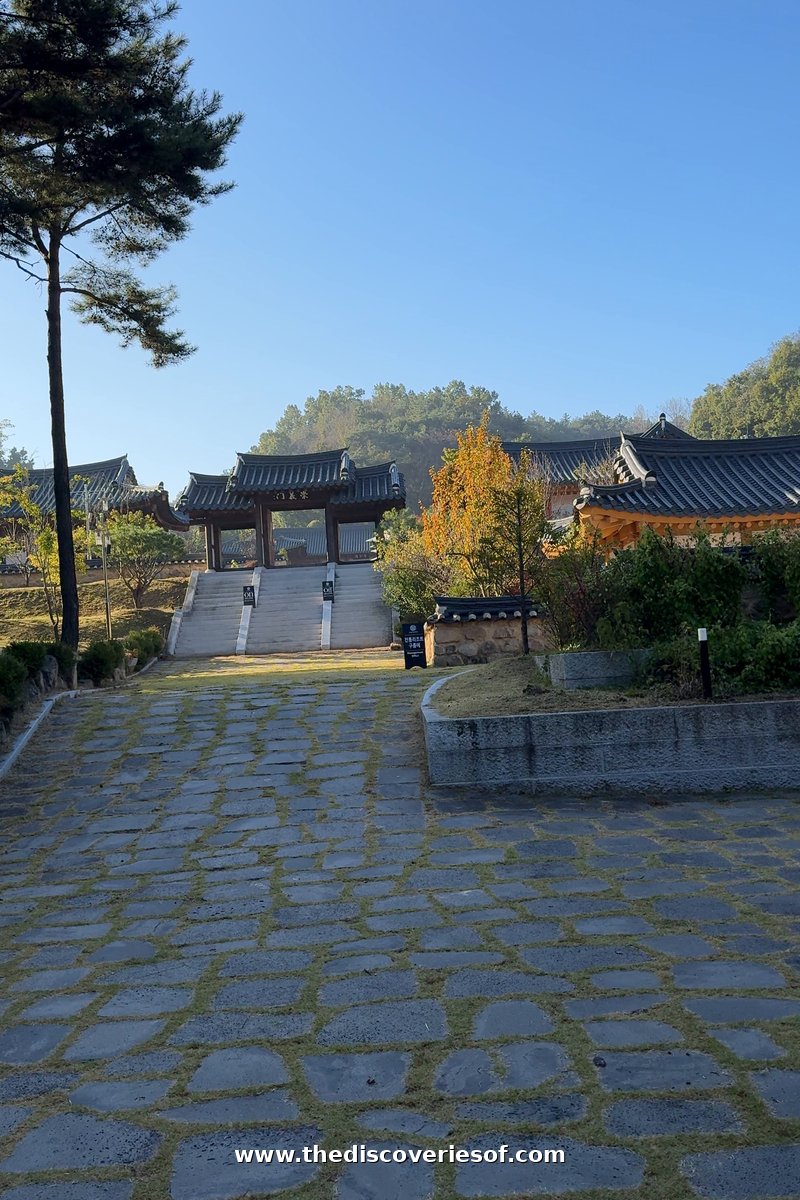 A stone-paved path leading up steps to a gatehouse and tiled hanok roofs under blue sky at Tradition Resort Gurume, Andong