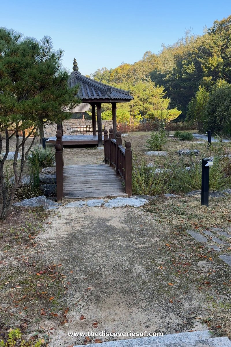 A wooden footbridge leading to a small tiled pavilion on a wooded hillside at Tradition Resort Gurume, Andong