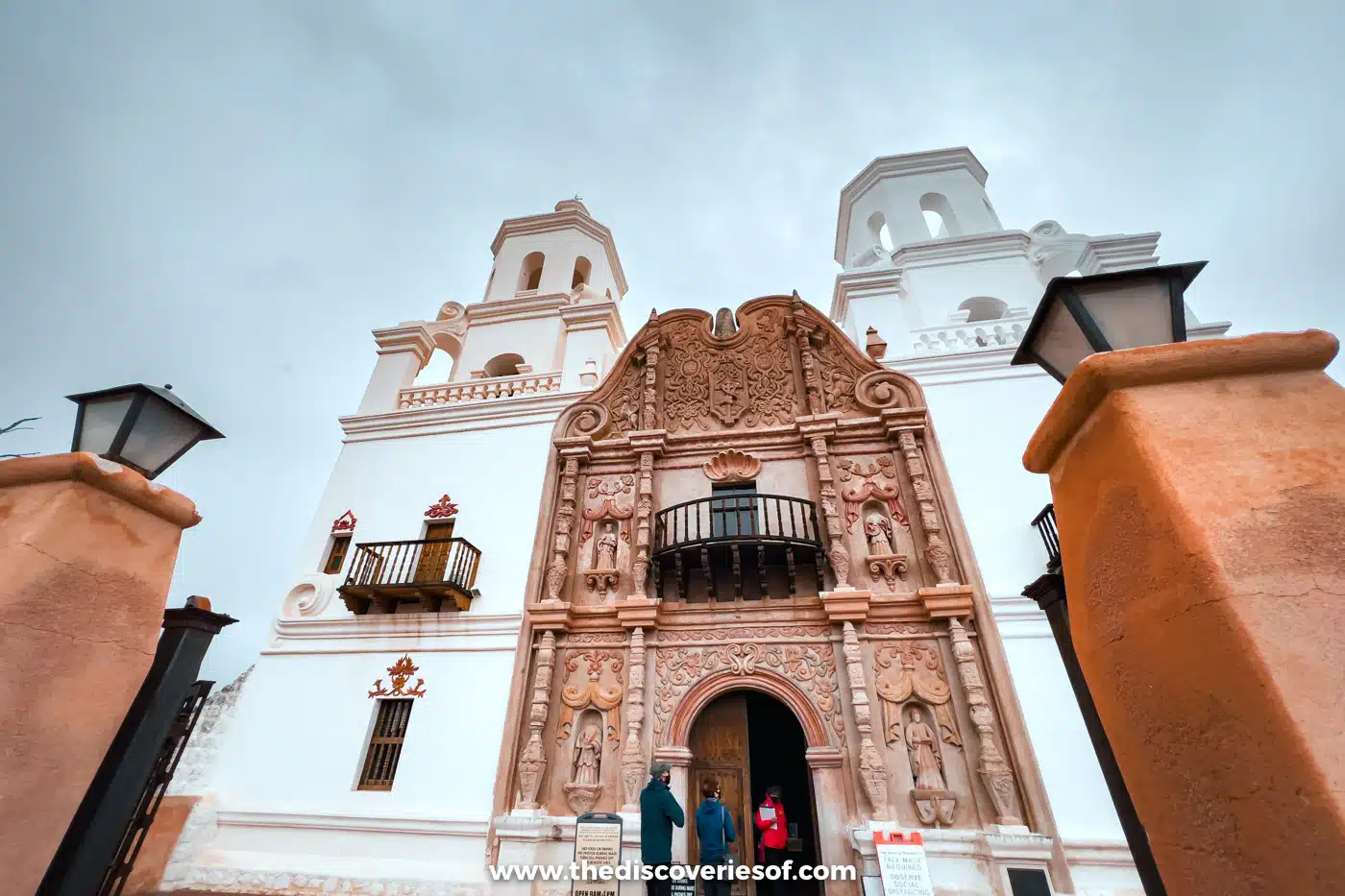 Looking up at the ornate carved entrance and white towers of Mission San Xavier del Bac between gateposts