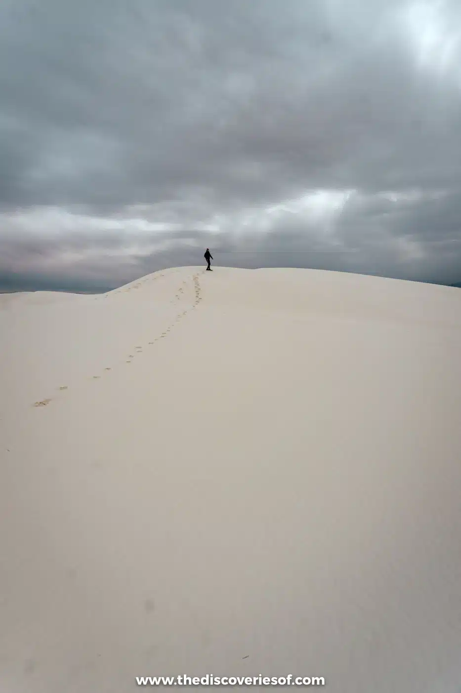 A single walker on the crest of a tall white gypsum dune under grey cloud at White Sands National Park