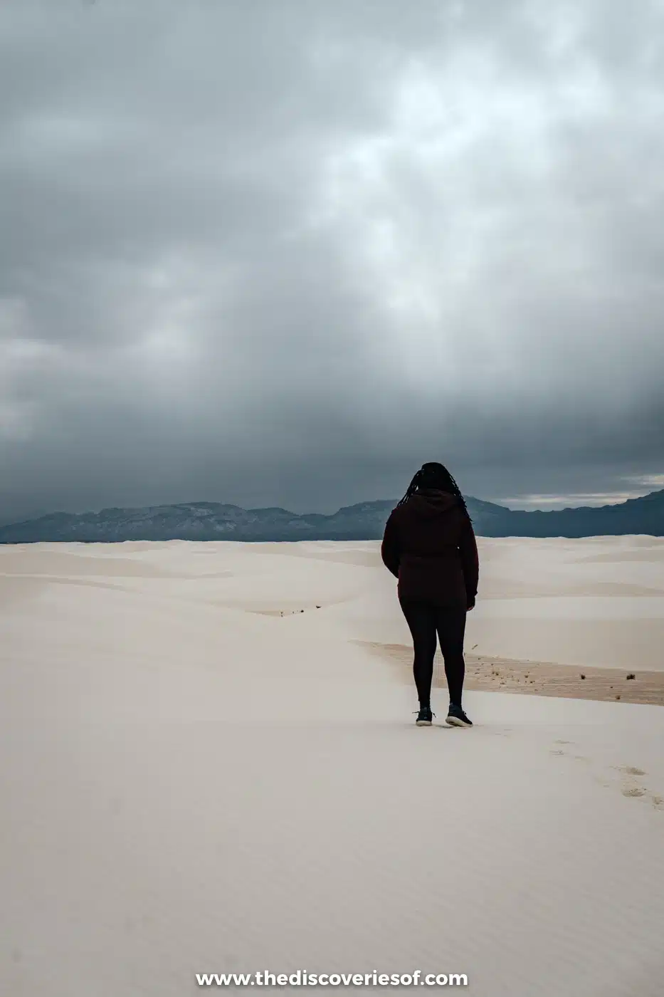 Woman in a dark coat standing alone on white gypsum dunes under heavy grey clouds at White Sands National Park