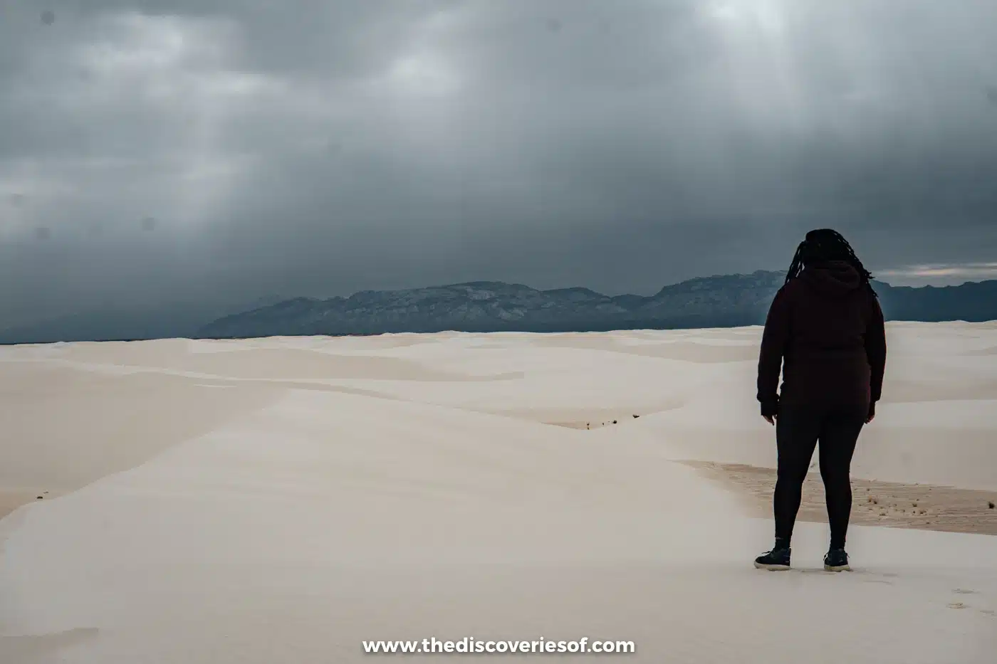 Julianna in a winter coat standing on the white gypsum dunes of White Sands National Park beneath dark rain clouds