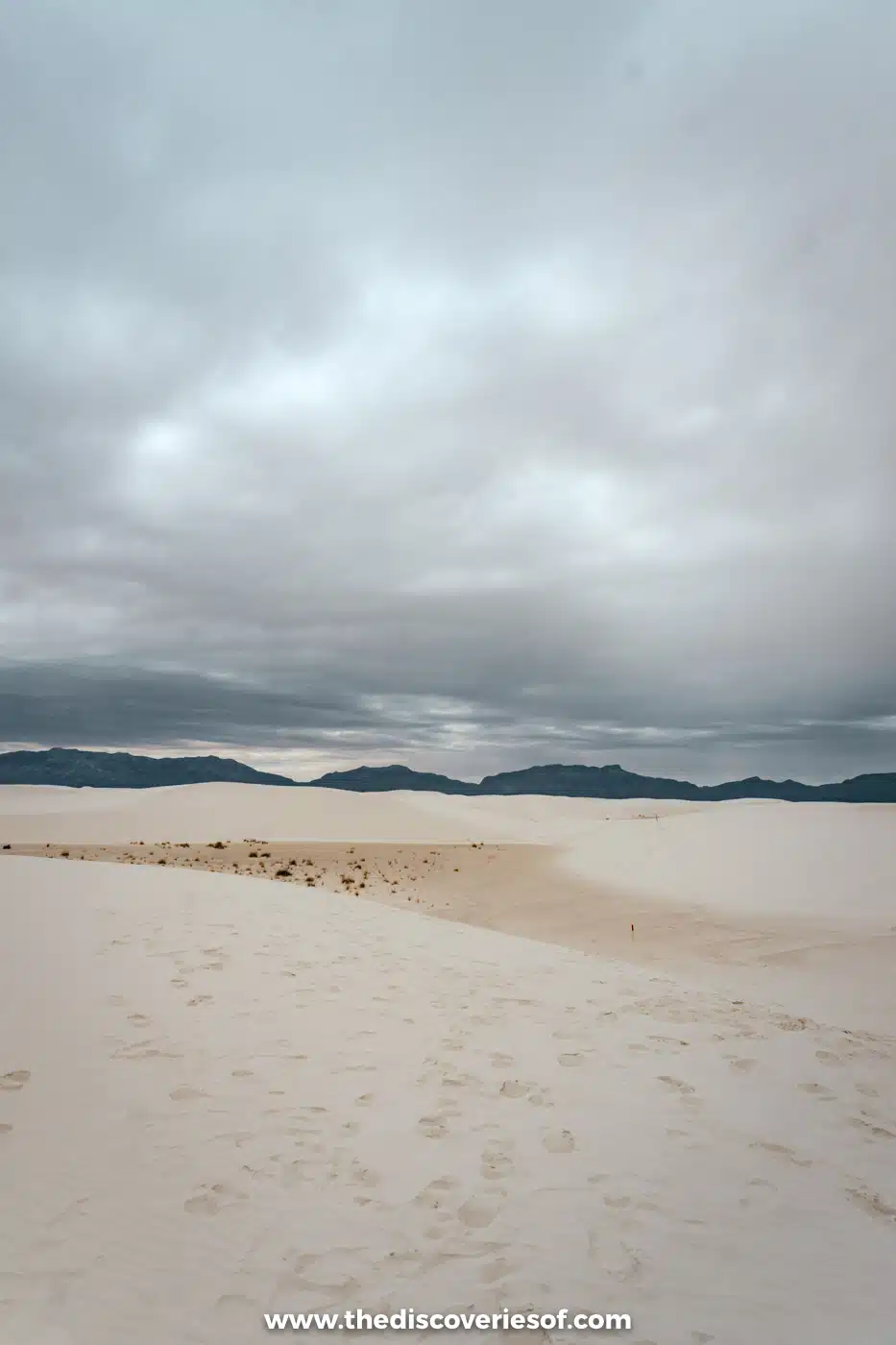 Footprints across white gypsum sand towards low dunes and mountains under grey cloud at White Sands National Park