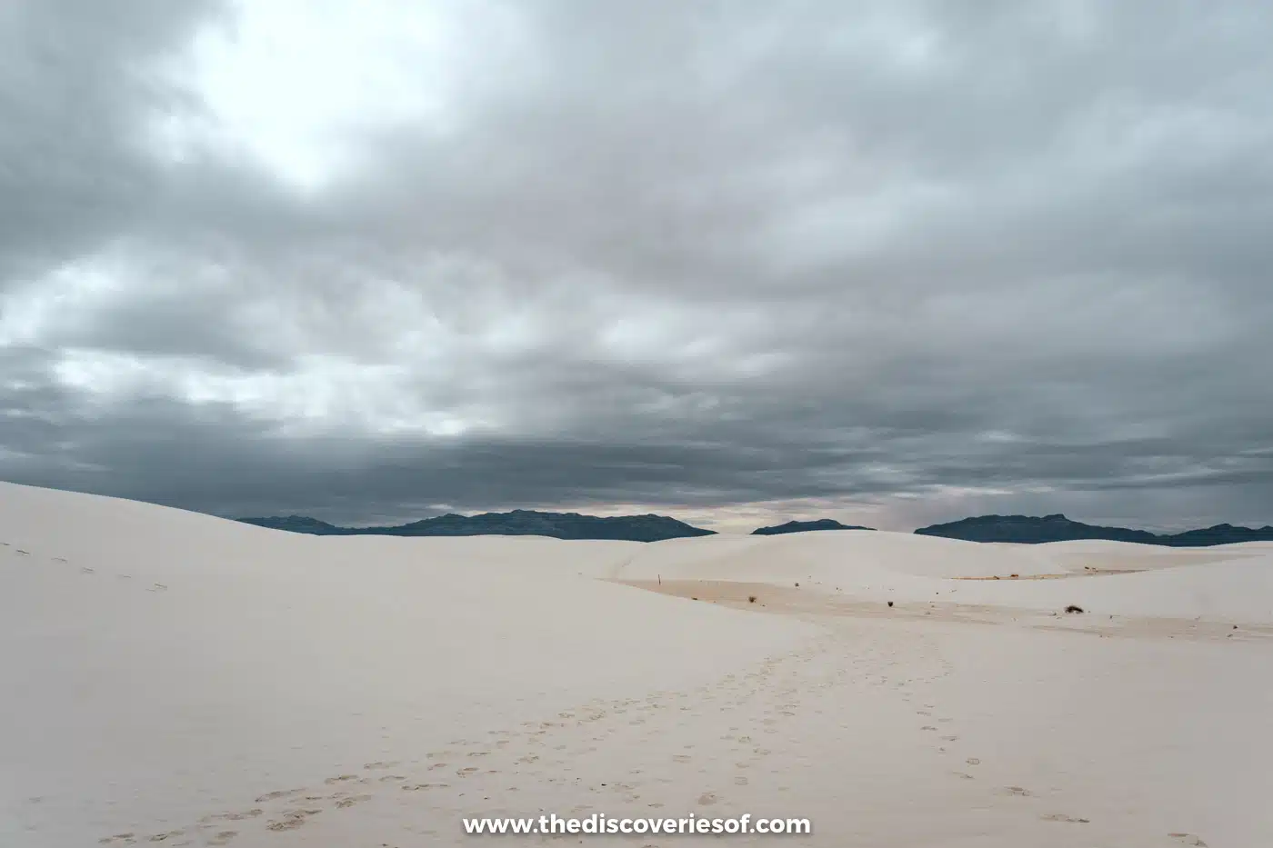 Footprints trailing across white gypsum dunes towards distant mountains under heavy cloud at White Sands National Park